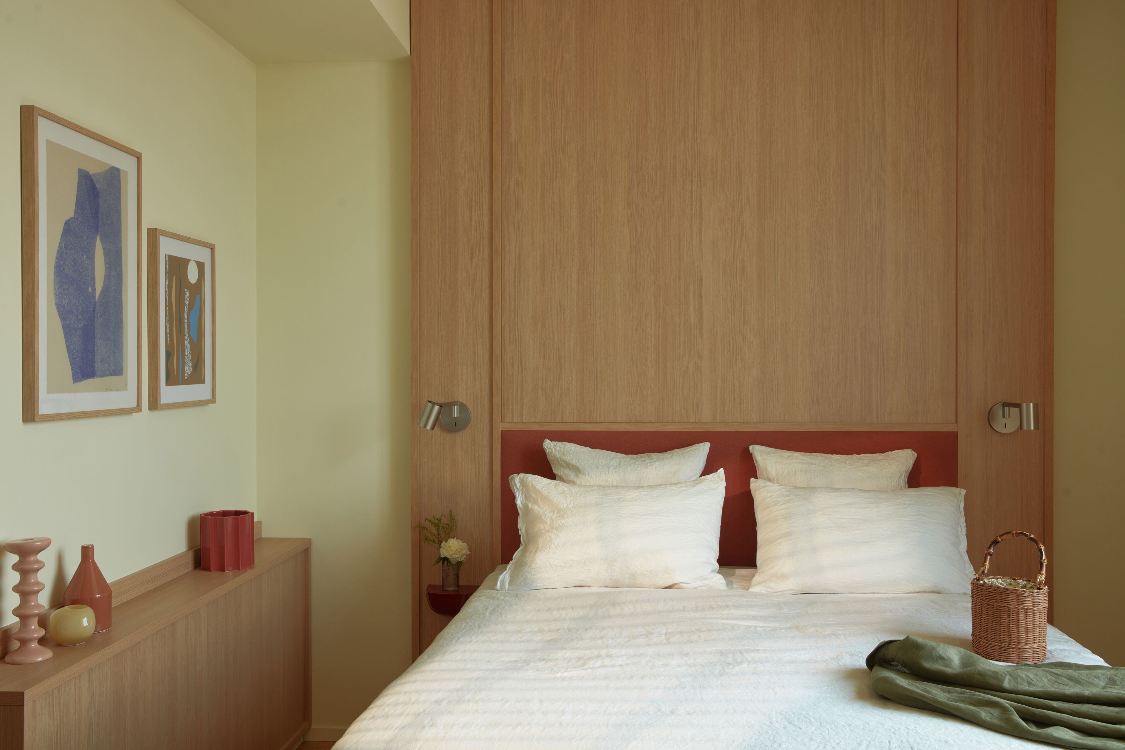 A neatly made bed with two pillows, a bedside table with a vase and a red container, and a wall-mounted clock.