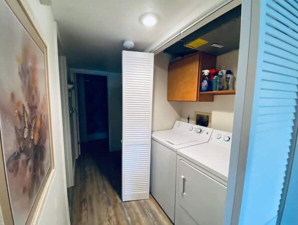 Un lavadero con lavadora y secadora, un armario de madera sobre la secadora, y una puerta que da a otra habitación.