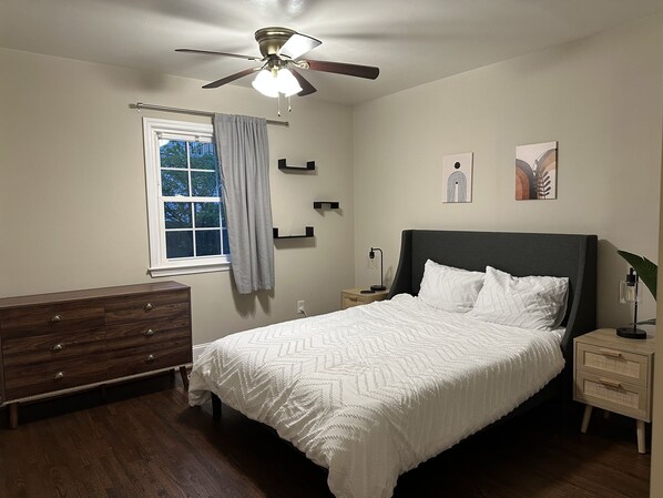 A bedroom with a bed, a dresser, a nightstand, a ceiling fan, a window with curtains, and wall art.