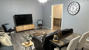 A modern living room with a black leather sofa, a dining table with chairs, a flat-screen TV, a clock, and a small plant on a wooden side table. - Modern 3BR 2BA West Enid (Enid)