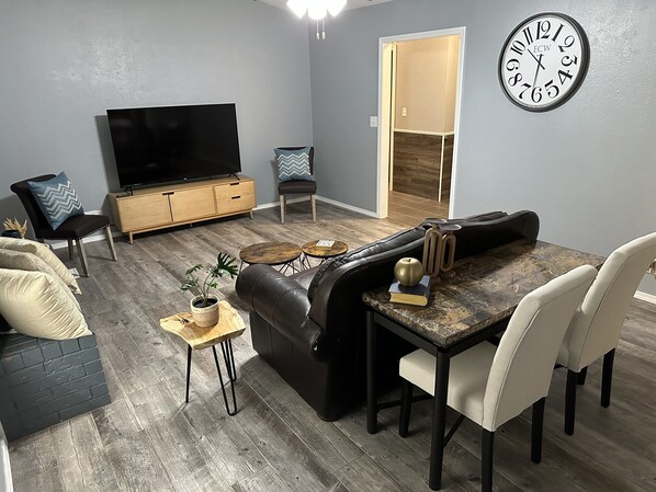 Una sala de estar moderna con un sofá de cuero negro, una mesa de comedor con sillas, un televisor de pantalla plana, un reloj y una pequeña planta sobre una mesita de madera.