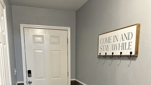 A hallway with a white door, a sign saying "COME IN AND STAY AWHILE", and dark wooden paneling on the wall. - Modern 3BR 2BA West Enid (Enid)