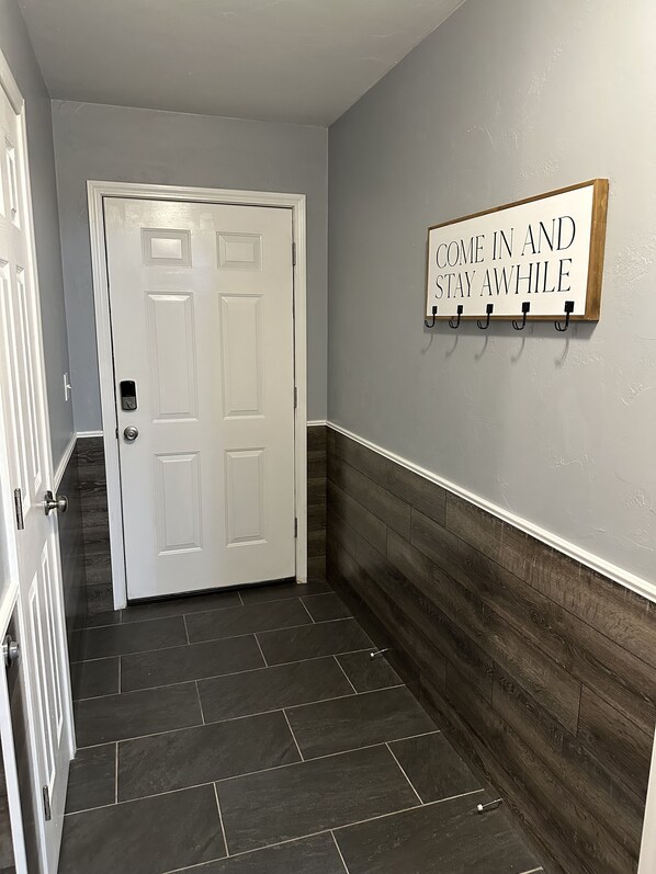 A hallway with a white door, a sign saying 'COME IN AND STAY AWHILE', and a wooden wall panel.