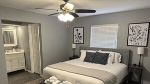 A bedroom with a bed, bedside tables, a nightstand, a ceiling fan, a window with blinds, a bathroom with a sink and mirror, and a door. - Modern 3BR 2BA West Enid (Enid)