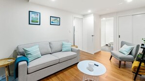 A modern living room with a grey sofa, a round coffee table, and a grey armchair. There are two framed pictures on the wall and a door leading to another room.