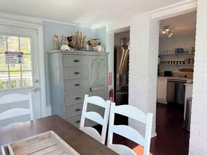 A dining area with a wooden table and white chairs, a grey cabinet with drawers, and a kitchen area with a stainless steel refrigerator and a water dispenser.