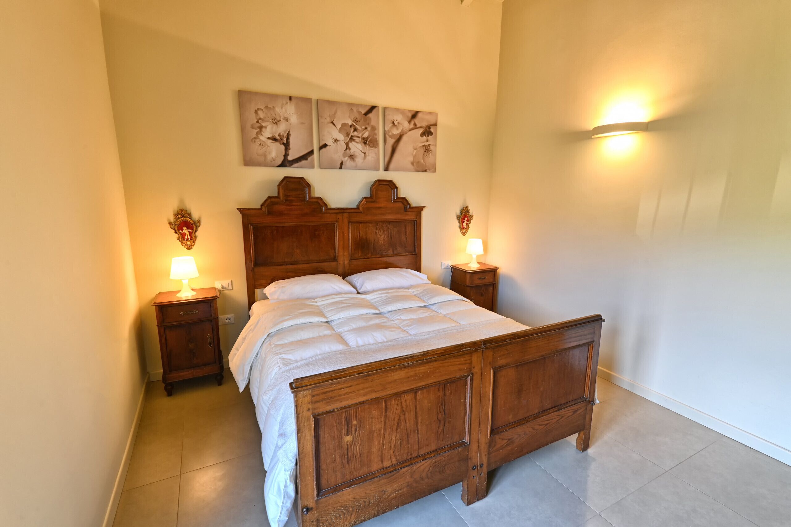 Uma cama de madeira com colcha, duas mesas de cabeceira com abajures e arandelas fixadas na parede.