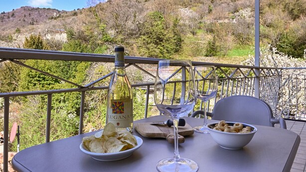 A table with a bottle of wine, glasses, chips, and a bowl of snacks on a balcony with a view of hills. - Appartamento Billy by Theater Home - with partial lake view and garage (Tignale)