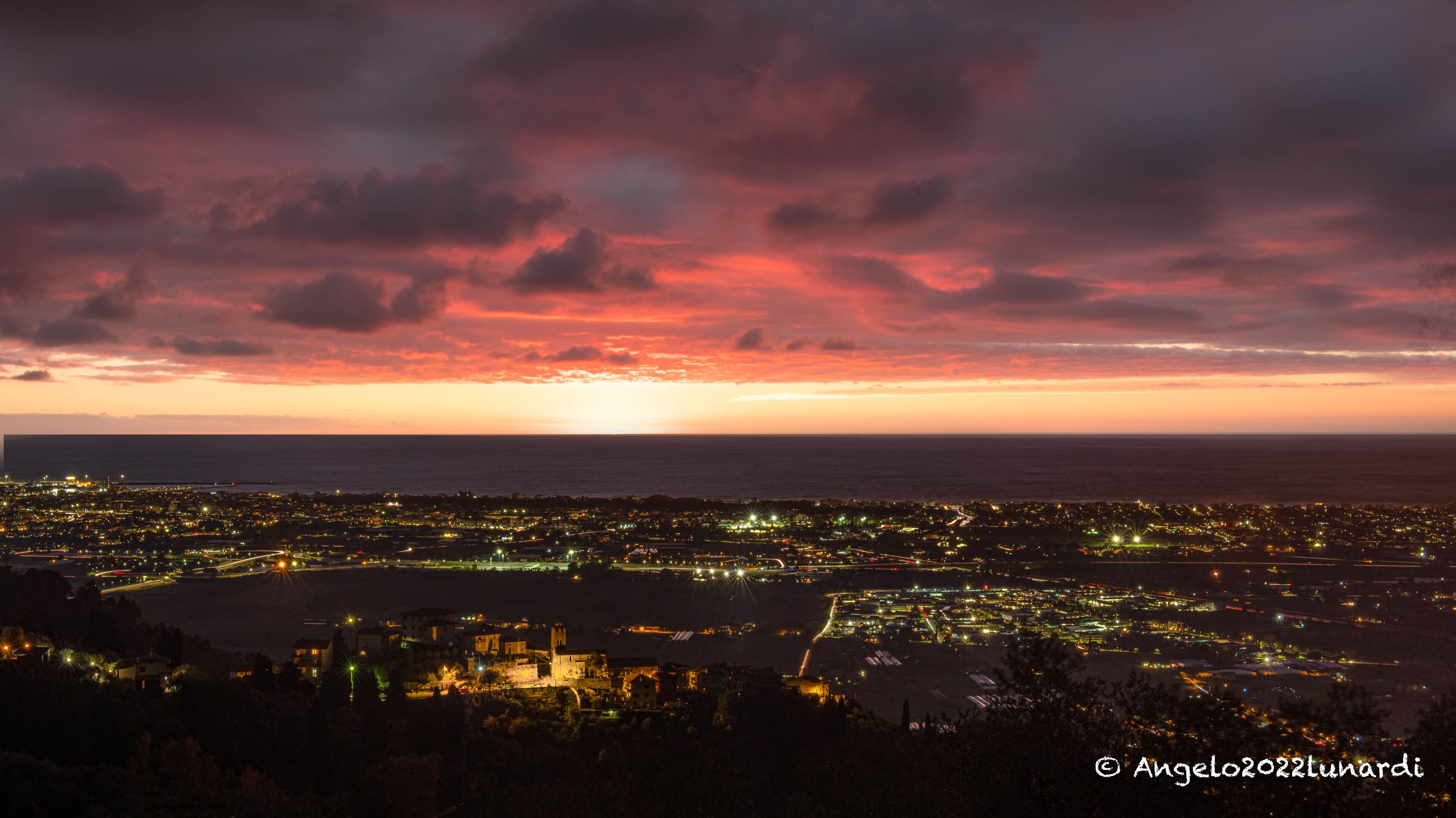 Un paisaje urbano al atardecer con el sol poniéndose sobre el océano, que muestra edificios iluminados y una vista despejada de la costa.