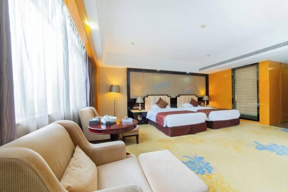 A hotel room with a sofa, a bed with two pillows, a bedside table with a lamp, and a large window with curtains.