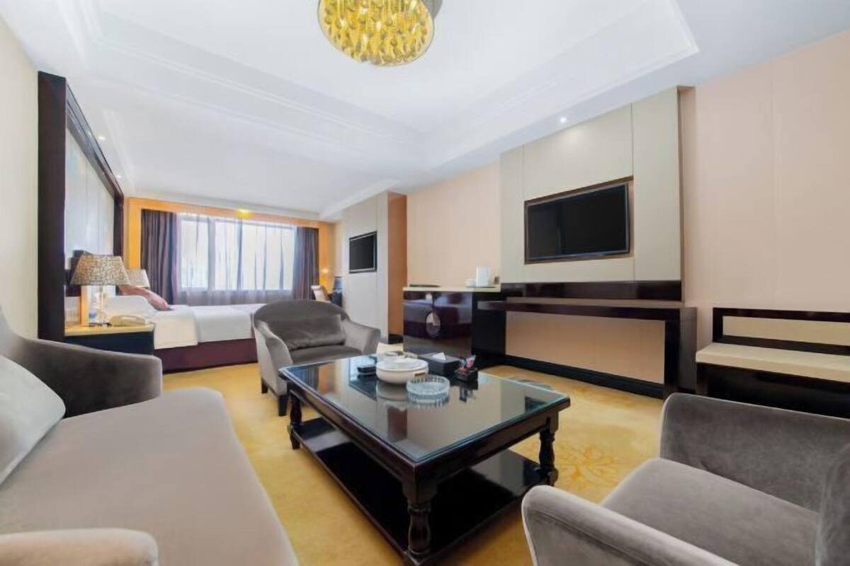 A hotel room with a sofa, armchair, a glass coffee table, a TV, and a large window.