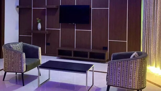 A modern living room with a flat-screen TV mounted on a wooden panel wall, two patterned armchairs, a coffee table, and ambient blue lighting. - Debbie's Suites and Apartments  (Orozo)