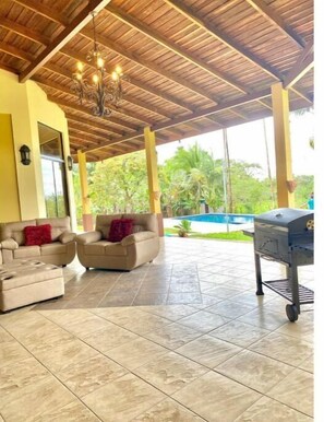 An outdoor patio with a wooden ceiling, two beige sofas with red cushions, a grill, and a view of a pool and greenery.