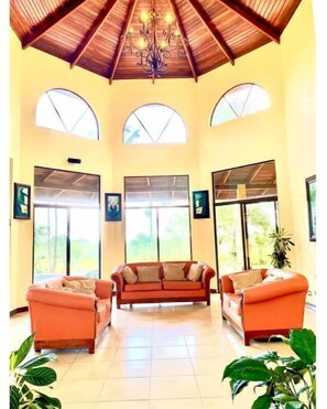 A spacious room with a high ceiling, a chandelier, and large windows. There are three sofas and two potted plants.
