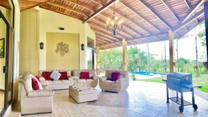 Un espacio de estar al aire libre techado con dos sofás y una mesa de centro, una parrilla y una piscina al fondo.