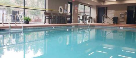 A clear indoor swimming pool with a lifeguard chair, pool ladder, and a view of the outside through large windows.