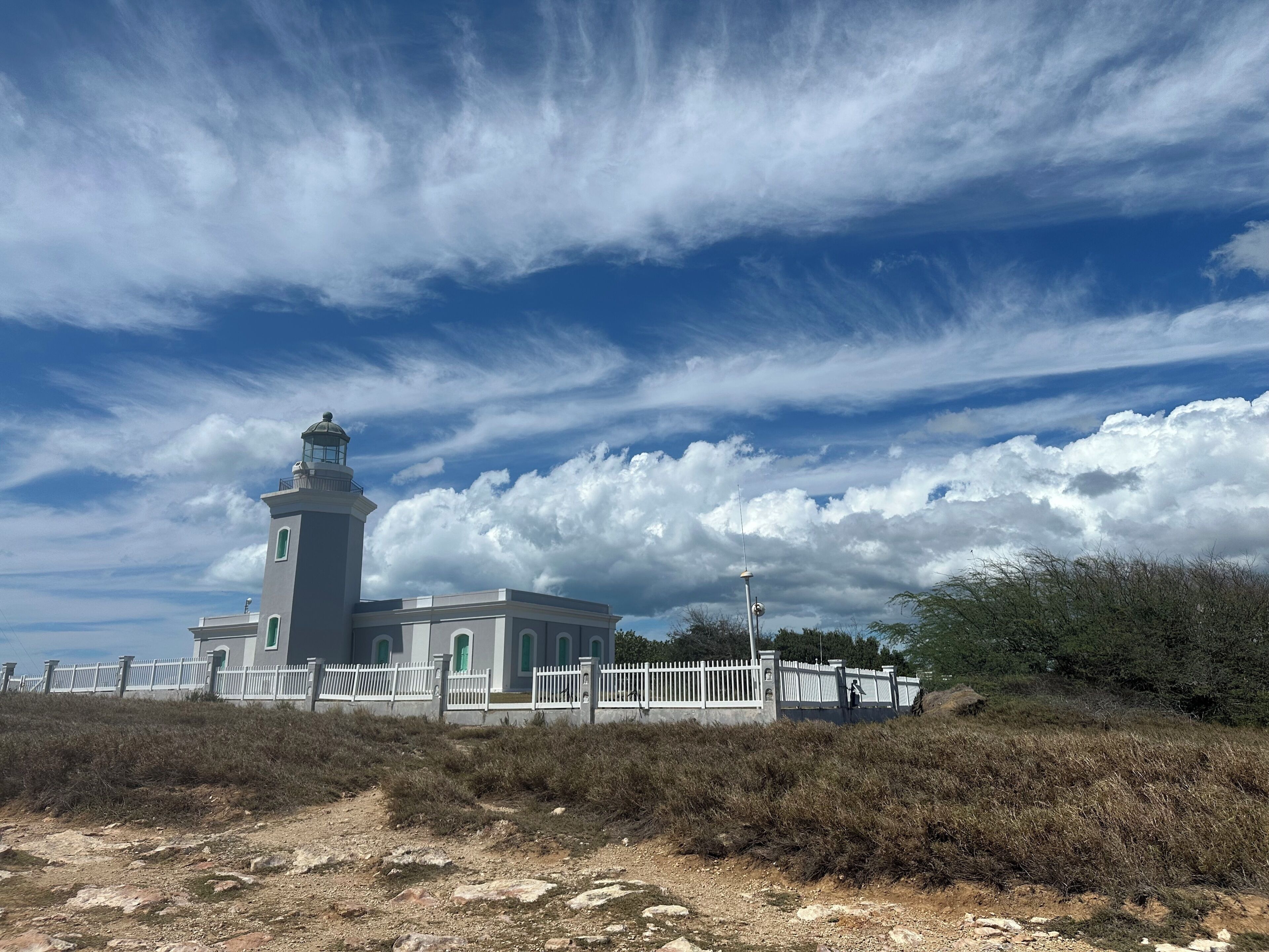 A lighthouse with a white fence, a grassy area, and a clear sky with clouds.