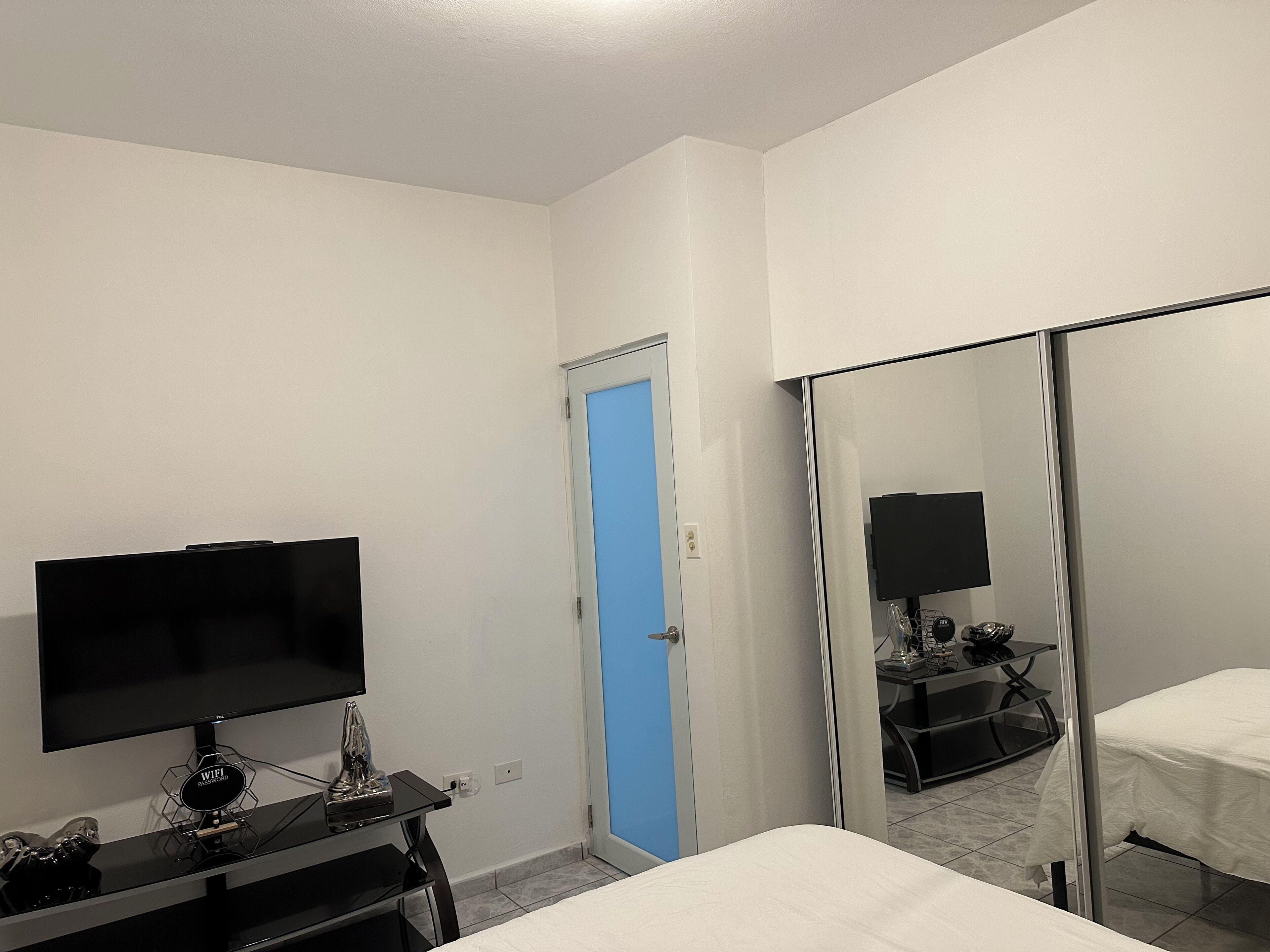 Habitación de hotel con cama, televisión, escritorio con computadora, espejo y puerta.