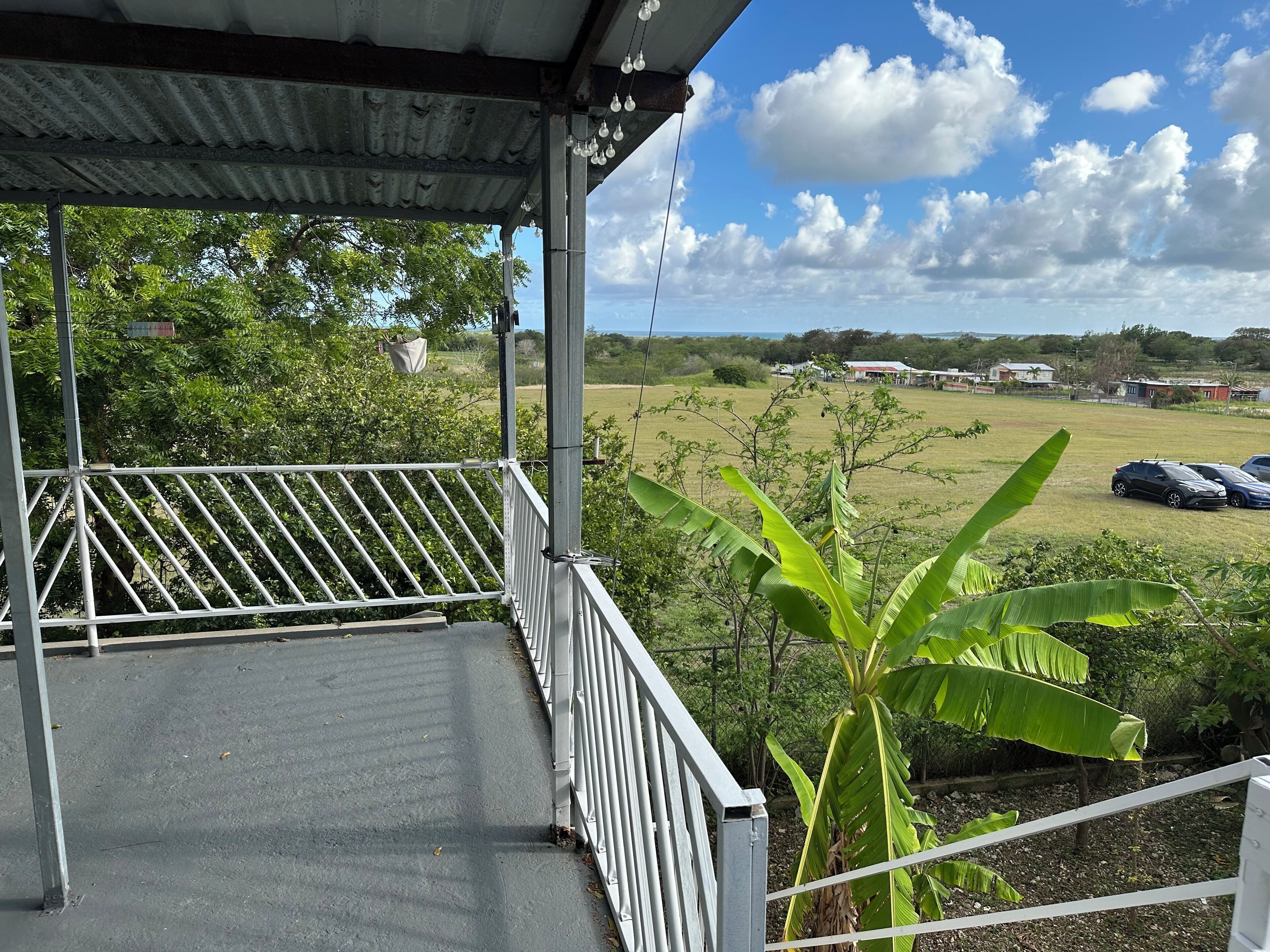 A covered outdoor area with a metal railing, a concrete floor, and a view of a grassy field with trees and buildings in the distance.