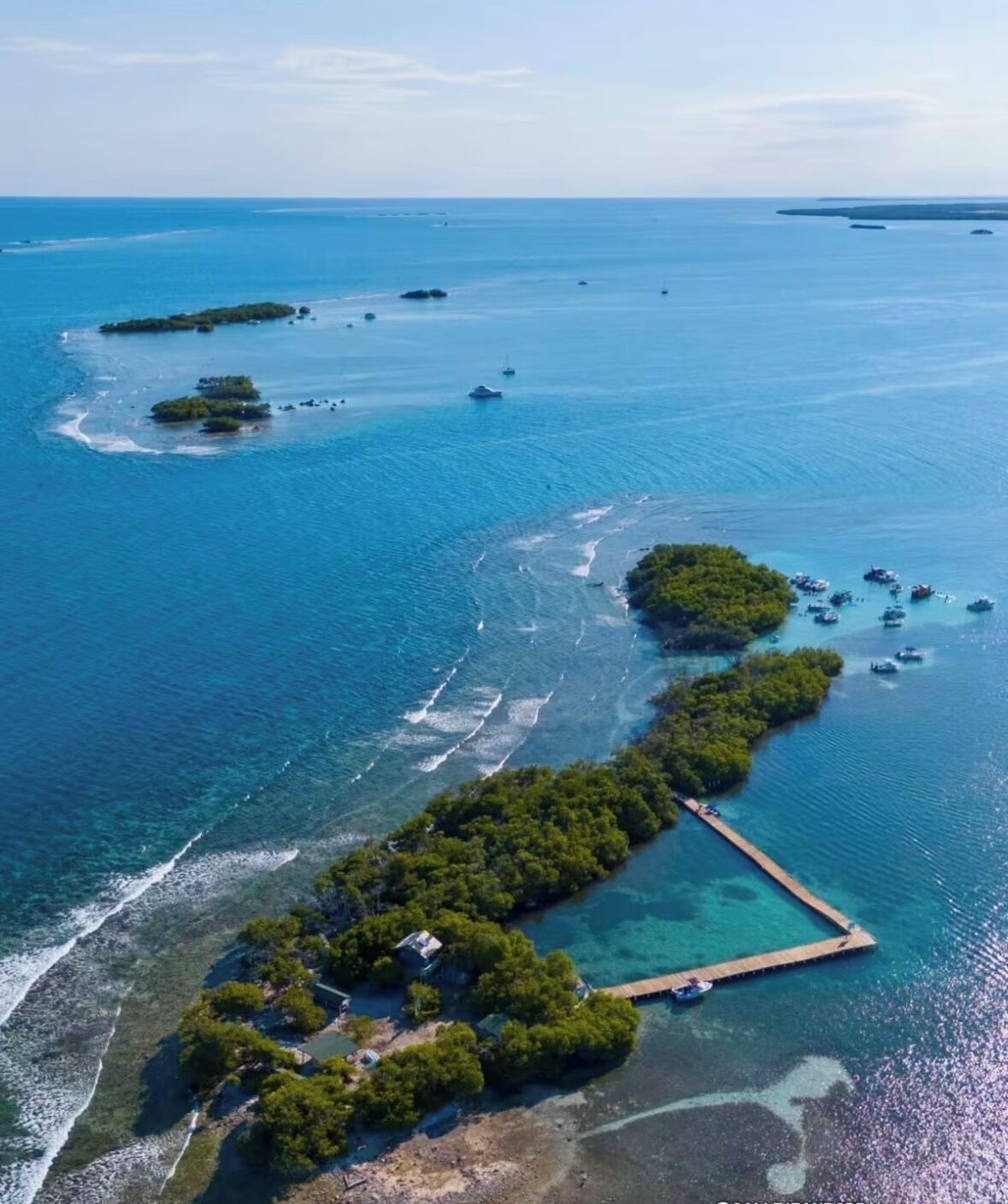 Vue aérienne d’une île tropicale aux eaux d’un bleu limpide, avec une jetée et plusieurs bateaux.