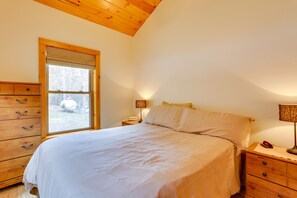 Ein Schlafzimmer mit hölzernem Kopfteil, einem großen Bett mit weißer Bettwäsche, einer Kommode mit Schubladen, einem Nachttisch mit Lampe und einem Fenster mit Blick ins Freie.