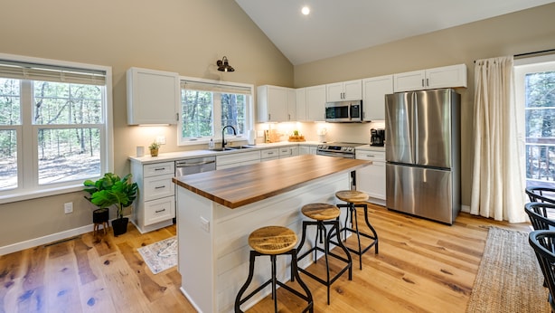 A modern kitchen with white cabinets, a stainless steel refrigerator, a wooden island with stools, and large windows offering a view of trees. - Chic Wisconsin Retreat w/ Deck, Grill & Fire Pit! (Wisconsin Dells)