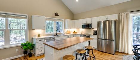 A modern kitchen with white cabinets, a stainless steel refrigerator, a wooden island with stools, and large windows offering a view of trees.