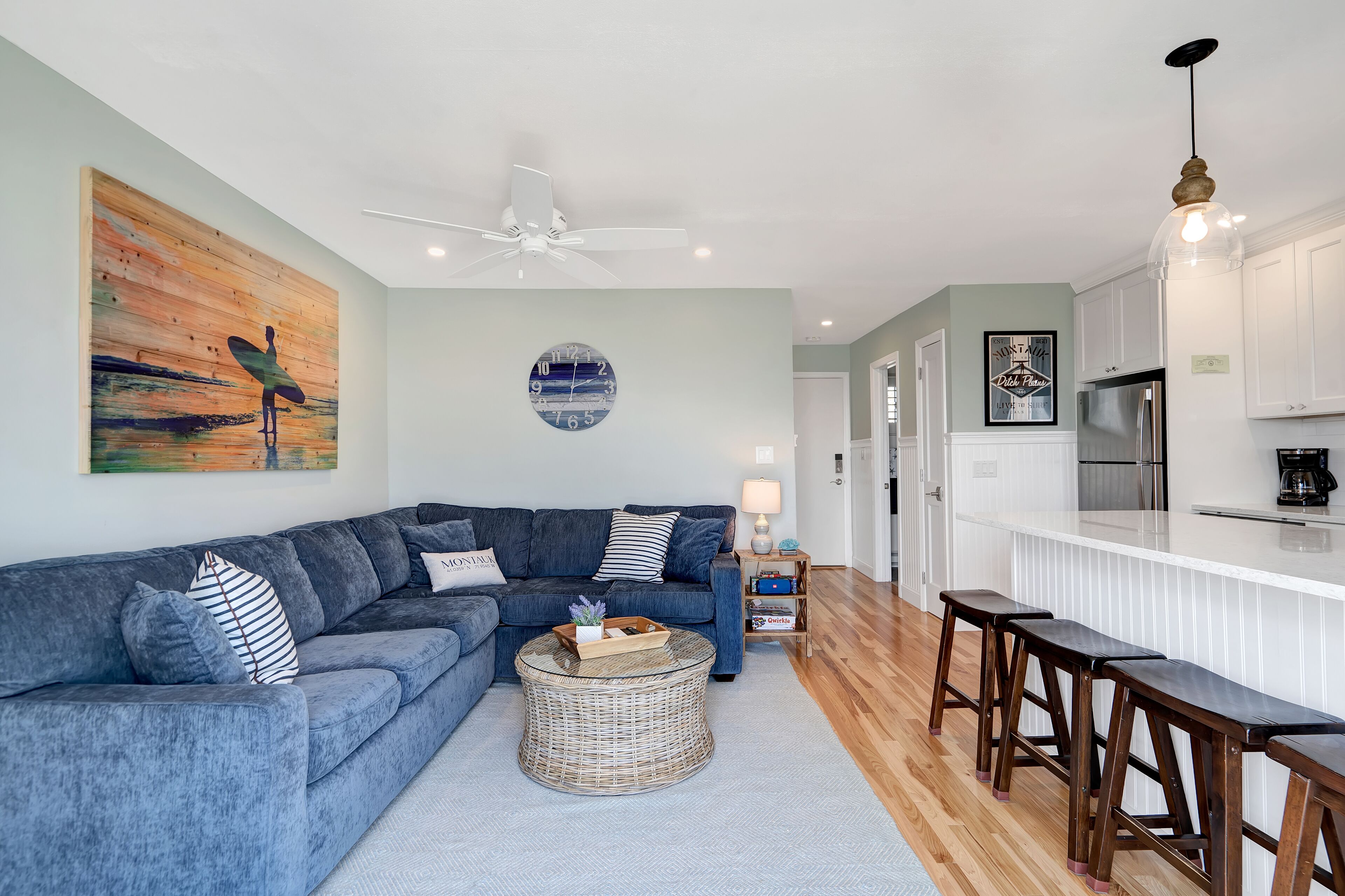A modern living room with a blue sectional sofa, a round wicker coffee table, and a kitchen area with white cabinets and a stainless steel refrigerator.