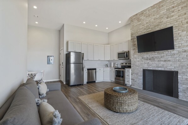 A modern kitchen with white cabinets, stainless steel appliances, and a flat-screen TV mounted on a brick wall. - Atlantic Bluffs Club (Montauk)