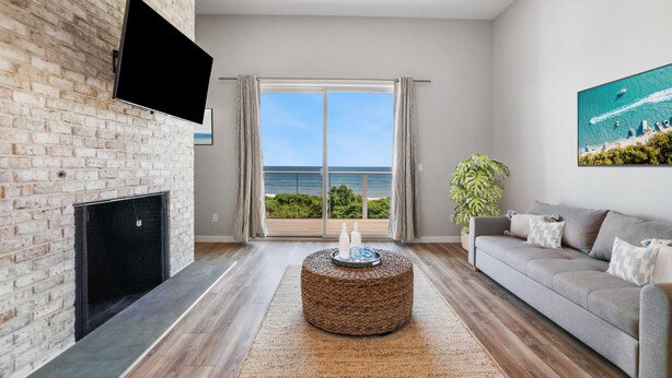 A modern living room with a fireplace, a flat-screen TV mounted on a brick wall, a grey sofa, and a large window offering a view of the ocean. - Atlantic Bluffs Club (Montauk)