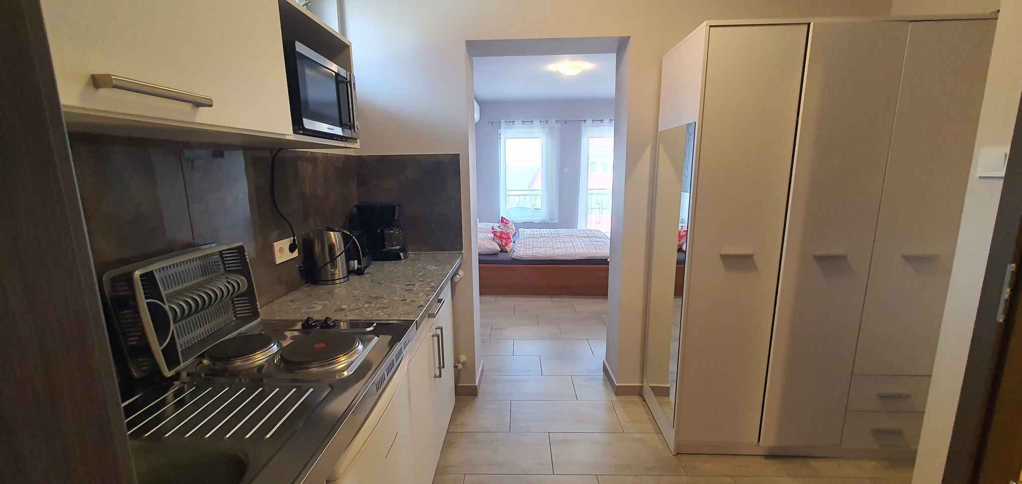 A narrow hallway with a kitchen area on the left, featuring a stove, microwave, and coffee maker. A bed with a white and red bedspread is visible in the background, leading to a room with a window.