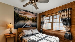 Un dormitorio con ventilador de techo de madera, cama con colcha, mesita de noche con lámpara, ventana grande con cortinas y un mural de un lago.