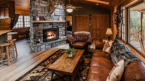 Interior acogedor de una cabaña con chimenea de piedra, muebles de cuero y un amplio ventanal con vistas al exterior.