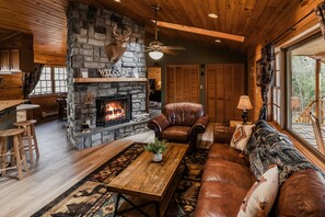 A cozy cabin interior with a stone fireplace, leather furniture, and a large window offering a view of the outdoors. - Catskills Cabin Rental Only 2.5 Hours Away From NYC! (Downsville)