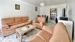 A living room with a beige sofa, a coffee table with a lace doily, a bookshelf, and a television on a stand. - Bright and quiet apartment near the sea (Saint-Raphaël)