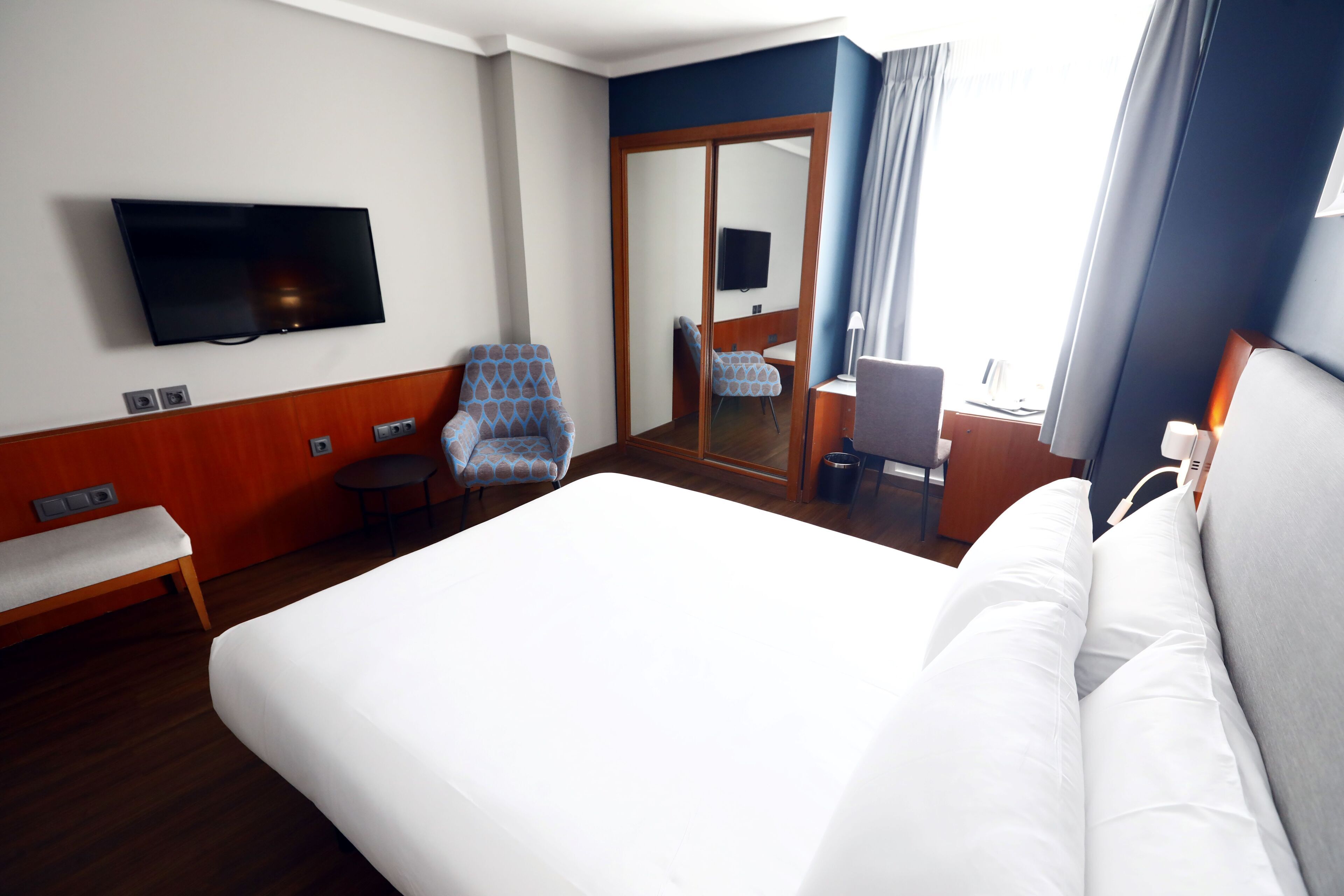 Habitación de hotel con una cama grande, un televisor montado en la pared, una silla, un escritorio y un espejo.