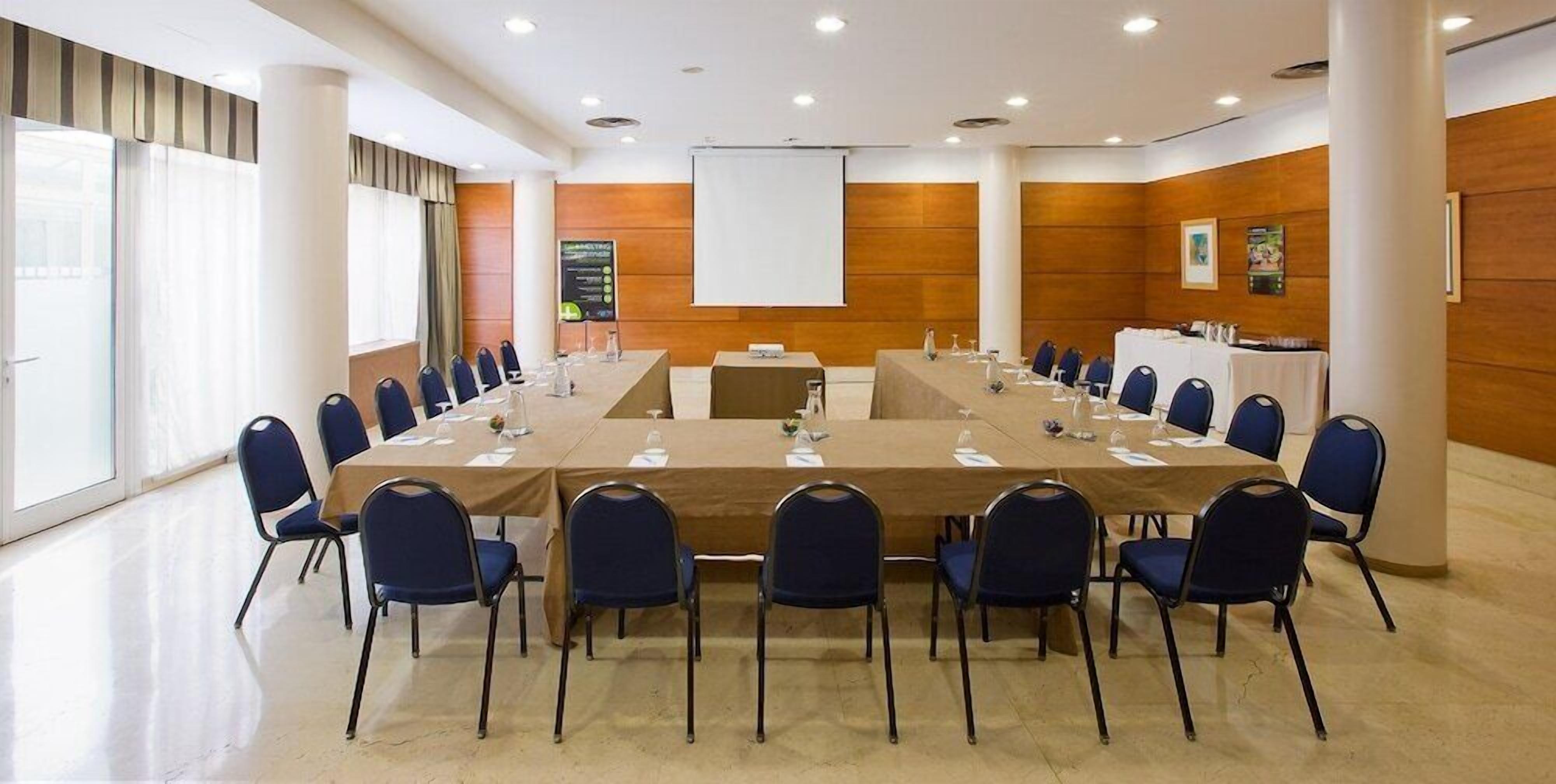 Ein Konferenzraum mit einem langen Tisch, blauen Stühlen und einer Projektionsleinwand.