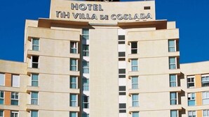 Un edificio de hotel con varios pisos, un letrero llamativo que dice "HOTEL VILLA DE COSLADA" y un cielo azul despejado al fondo.