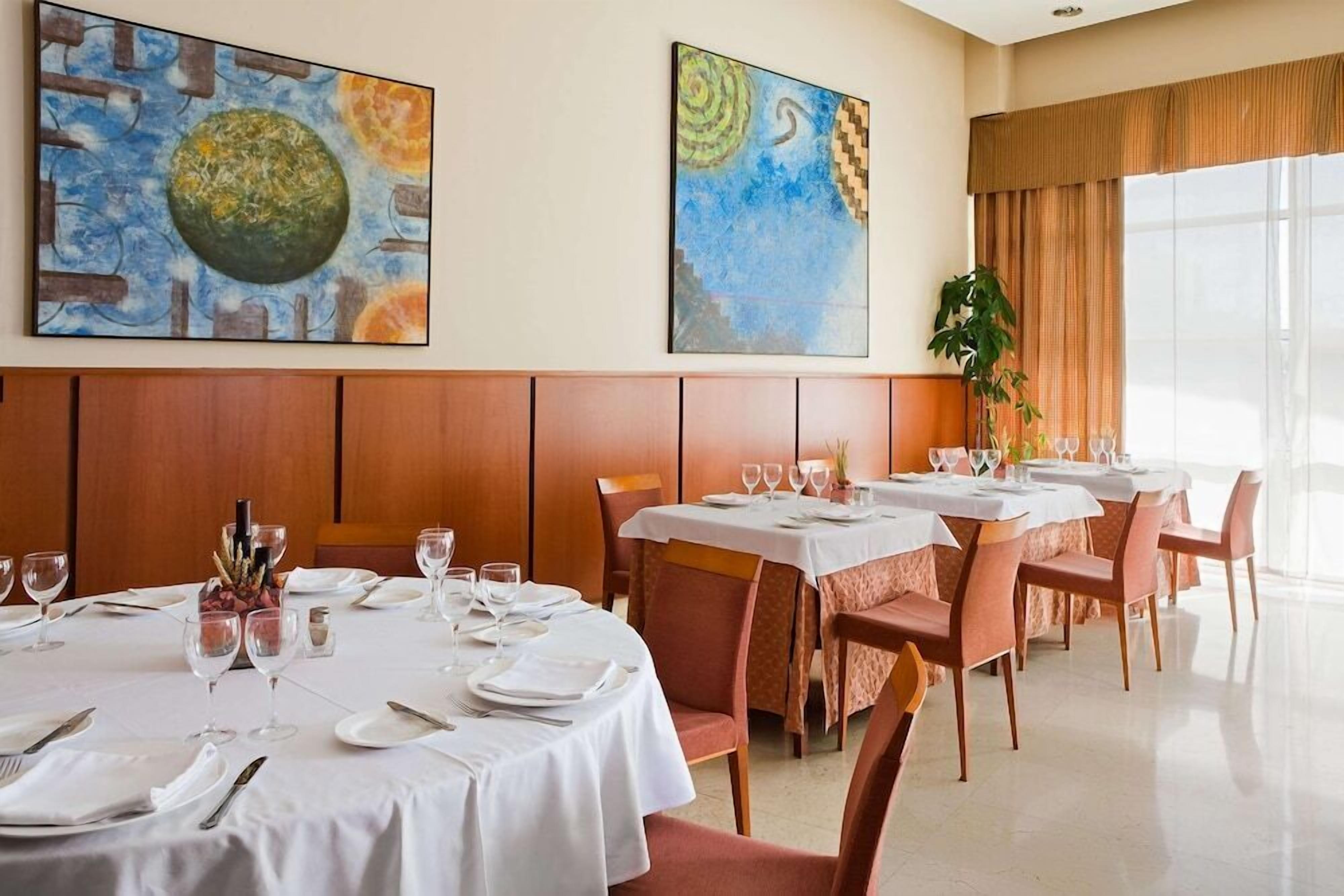 Comedor con mesas redondas para cuatro, manteles blancos y un ventanal con cortinas.