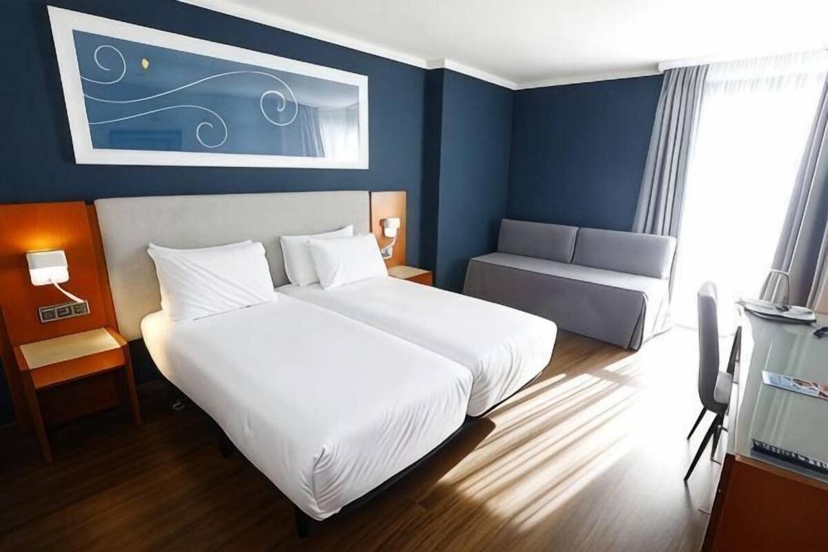 A hotel room with two beds, a sofa, a desk, and a large window.