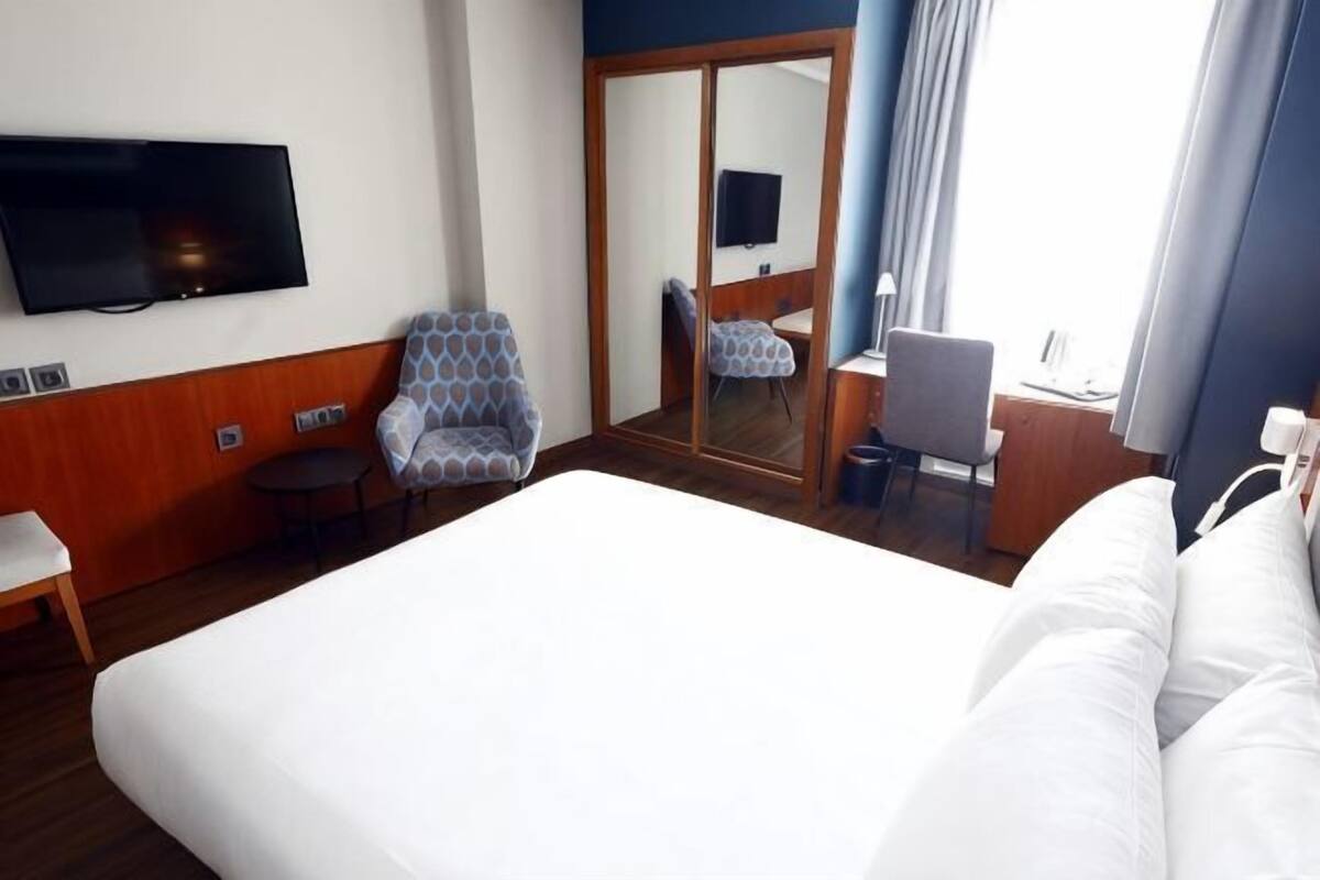 A hotel room with a large bed, a TV mounted on the wall, a seating area with chairs, and a mirror reflecting the room.