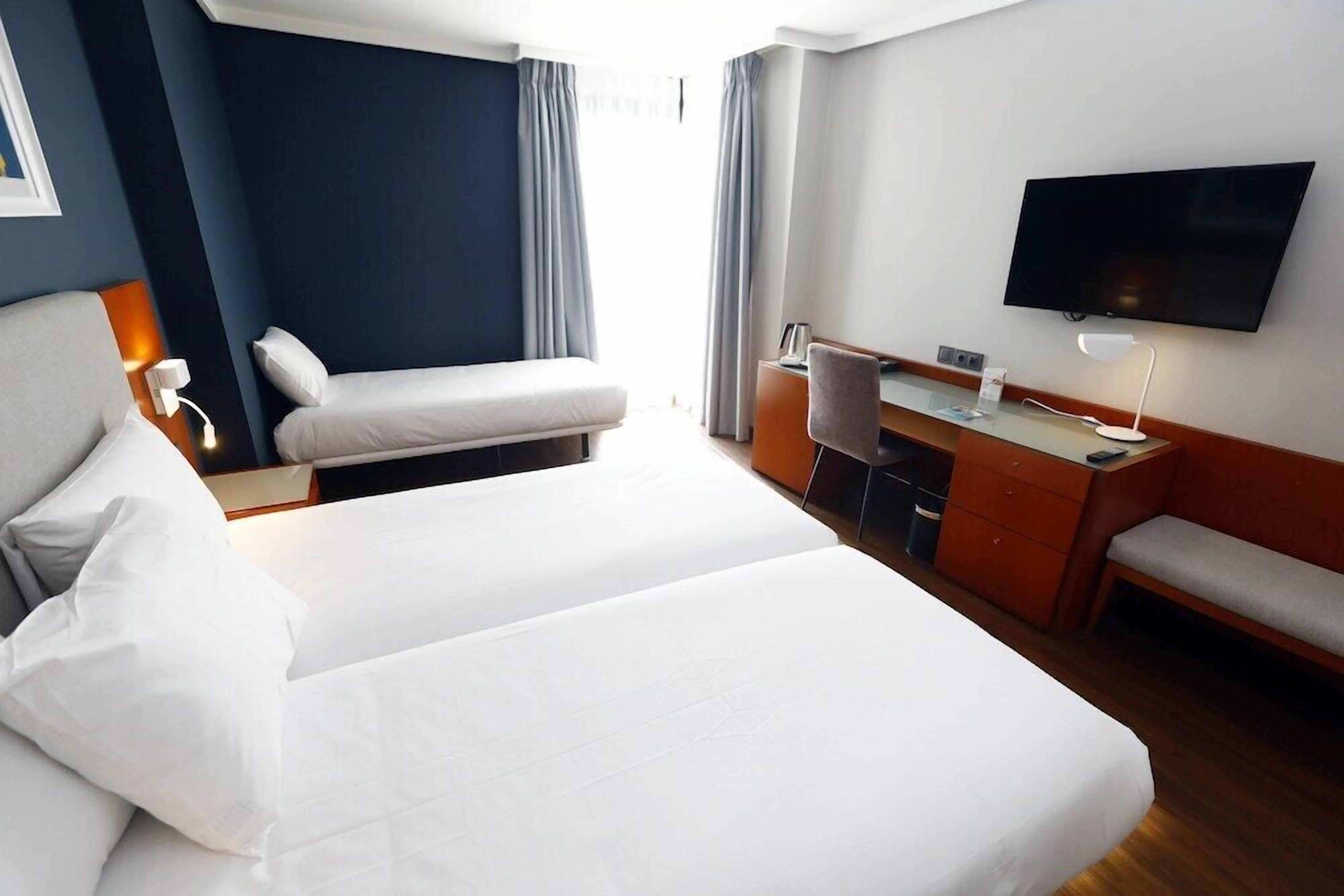 A hotel room with a large bed, a desk with a chair, a TV mounted on the wall, and a window with curtains.