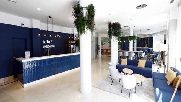 Un moderno vestíbulo de hotel con una recepción, zona de estar y plantas decorativas.