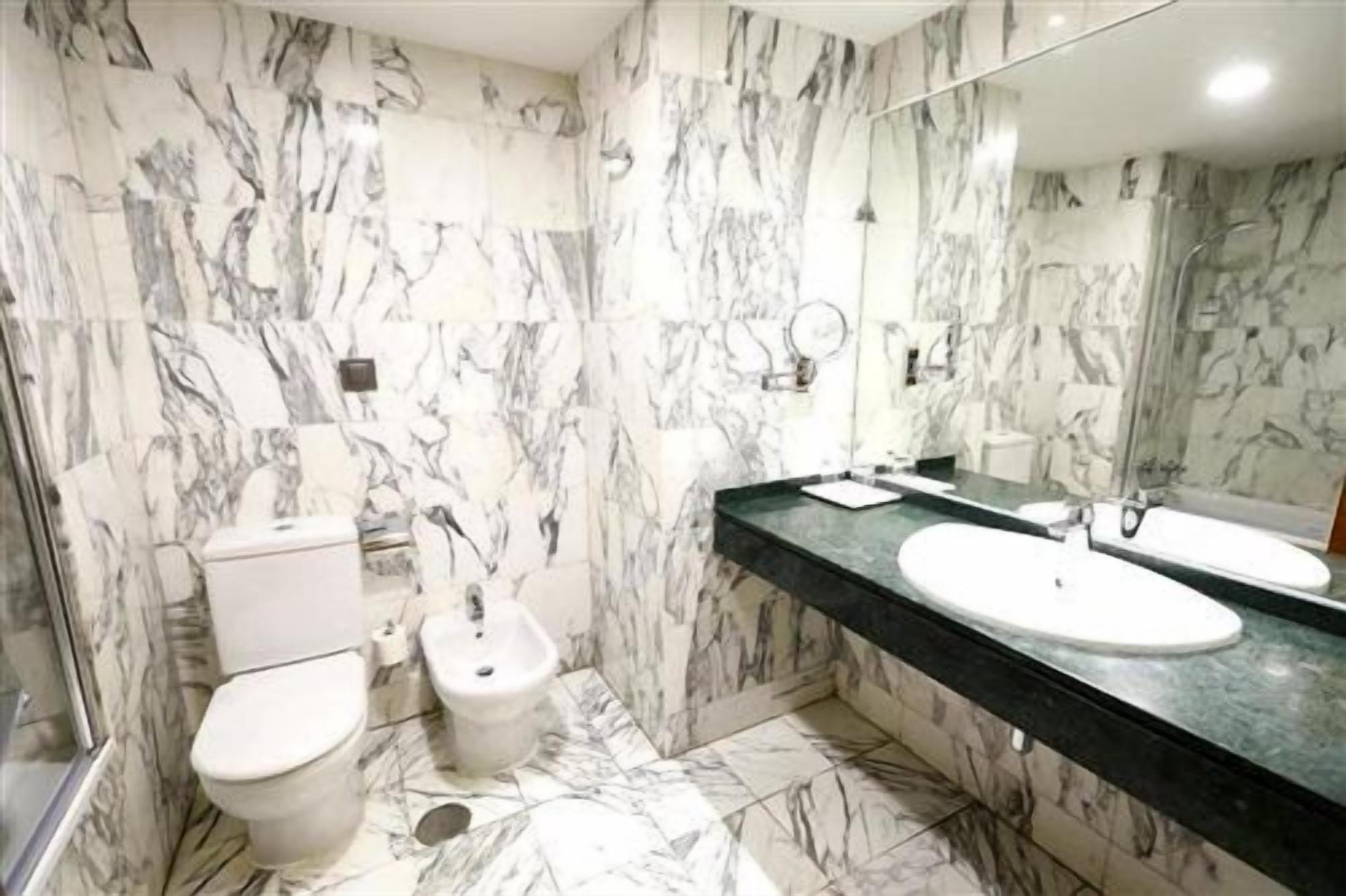 Une salle de bain avec des murs en marbre, des toilettes, un bidet, deux lavabos et un grand miroir.
