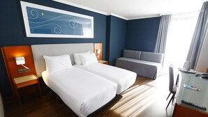 Habitación de hotel con dos camas, un escritorio, una silla, un sofá y una obra de arte en la pared.