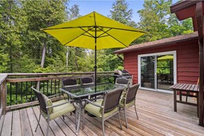 Een houten terras met een glazen tafel en stoelen, een gele parasol, en een rood gebouw met een schuifpui.