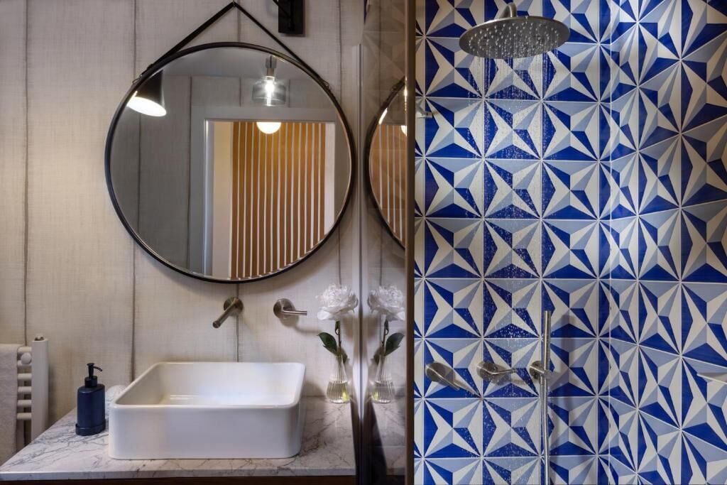 A bathroom with a white sink, a round mirror, and a geometric patterned shower wall.