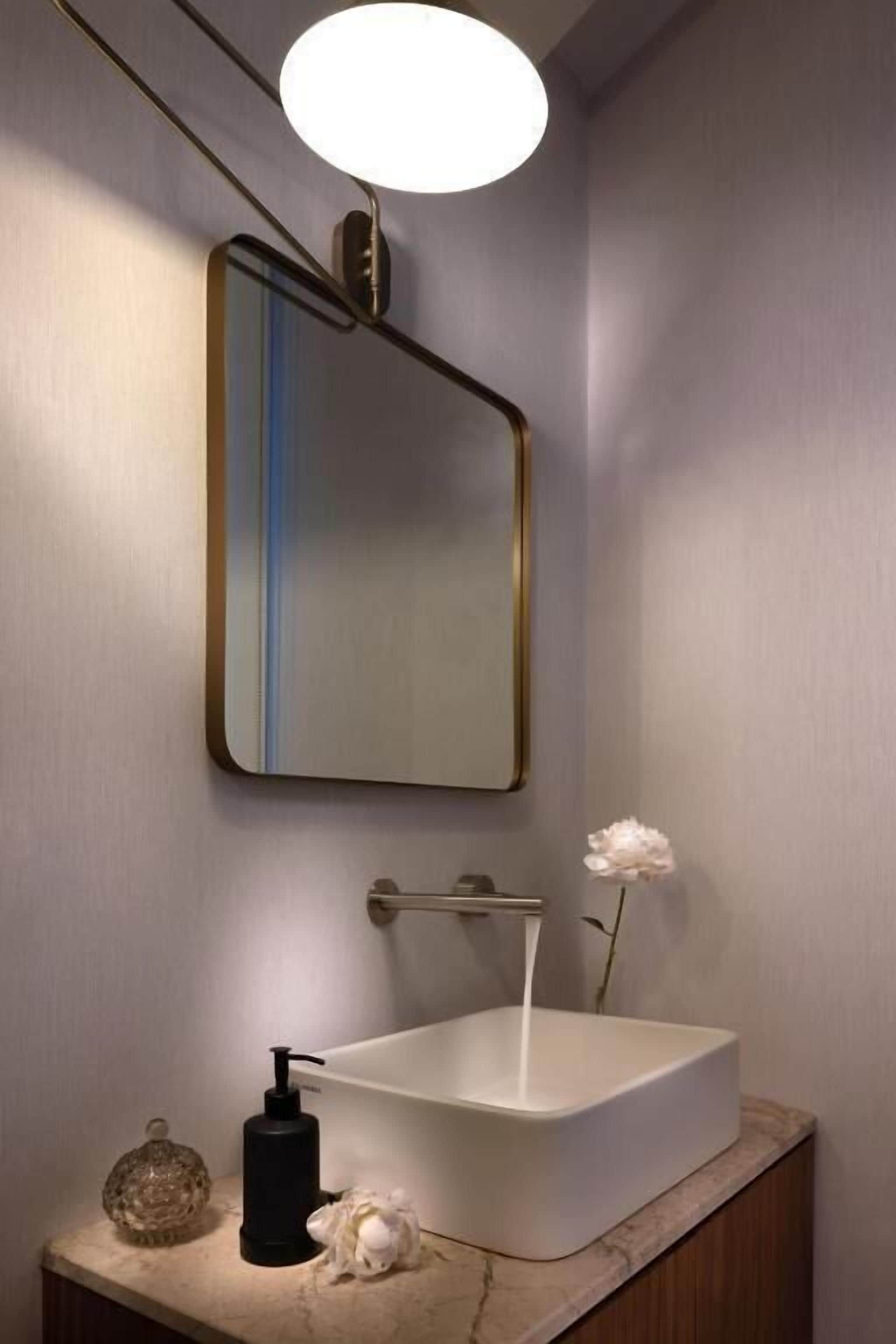 Ein Badezimmer mit einem weißen Waschbecken, einem Spiegel mit goldener Rahmung, einer runden Leuchte und einer Vase mit einer weißen Blume.