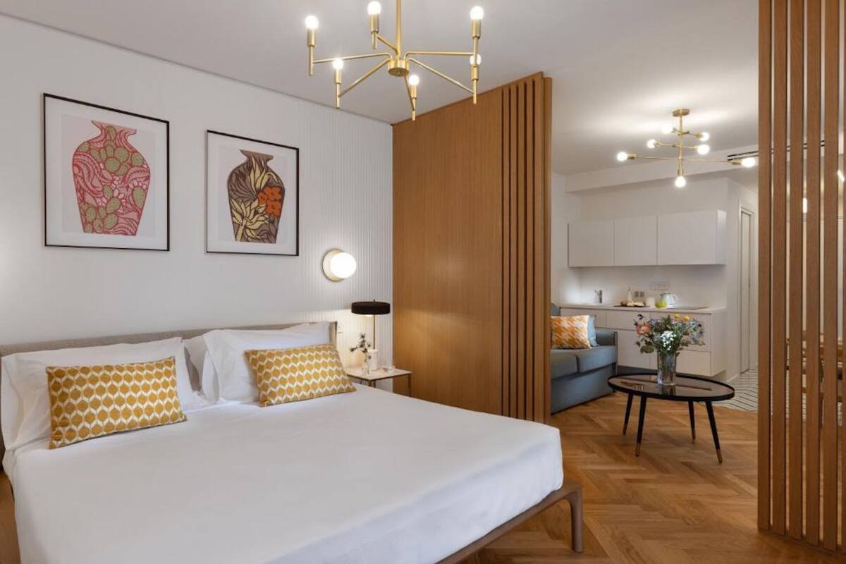 Una habitación de hotel moderna con una cama grande, una mesita de noche, una lámpara y una pequeña mesa redonda con un florero. Hay un separador de madera que divide la habitación de la zona de la cocina.