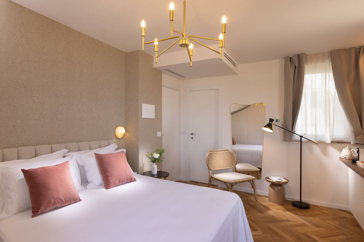 Una habitación de hotel con una cama grande, una mesita de noche con lámpara, una silla, una pequeña mesa con un jarrón de flores y un armario.